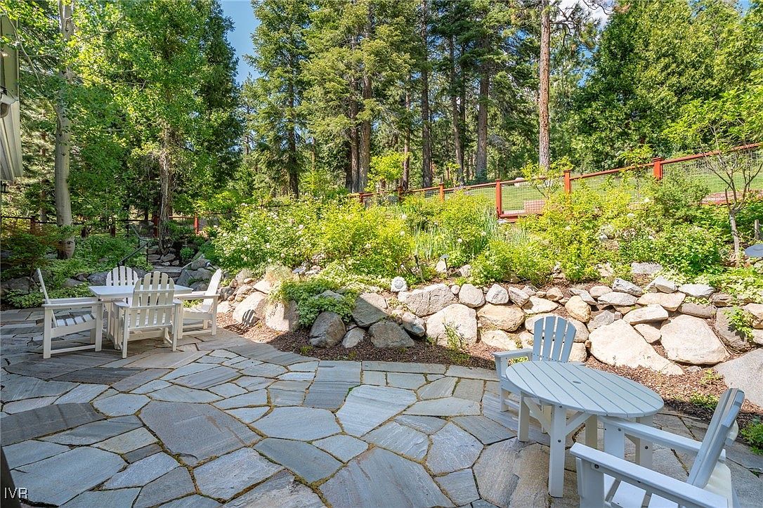 This image showcases a charming outdoor patio area, featuring a stone-paved surface and two sets of white patio furniture. The patio is surrounded by lush greenery, including trees, bushes, and a rock wall, creating a serene and private outdoor space. The scene evokes a sense of relaxation and outdoor living, perfect for entertaining or enjoying quiet moments.