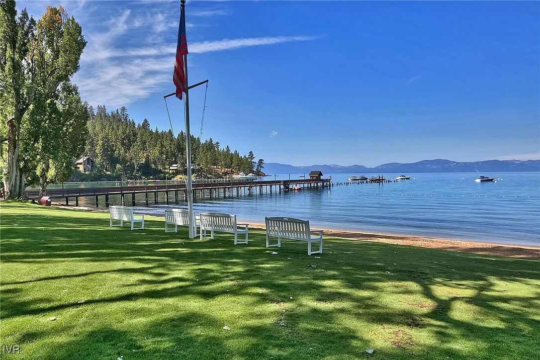 This exterior shot showcases a well-maintained yard with lush green grass leading to a sandy beach and calm blue water. Several white benches are arranged on the lawn, offering seating with a view of the lake and distant mountains. A pier extends into the water, adding to the scenic appeal of this lakeside property.