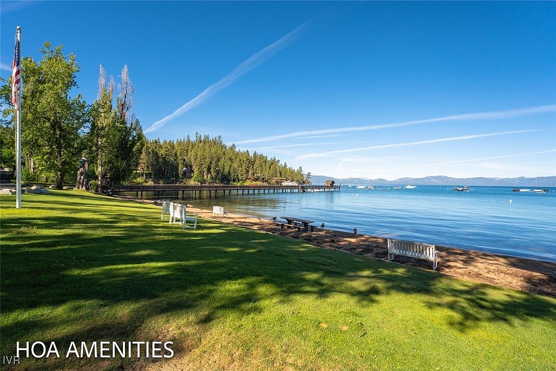 This image showcases a beautiful HOA amenity featuring a well-maintained grassy area leading to a serene lakefront. The scene includes benches and chairs for relaxation, a pier extending into the water, and a backdrop of lush trees and mountains. The overall impression is one of tranquility and upscale community living.