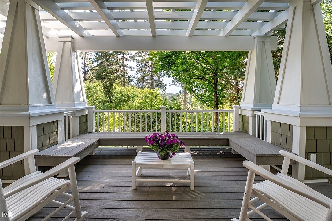 This image showcases a charming outdoor patio or deck area, featuring a white pergola overhead and built-in benches along the sides. A small white table with a potted plant sits in the center, adding a touch of nature. The deck is constructed with gray composite decking, and the view beyond the railing reveals lush greenery and trees, creating a serene and inviting atmosphere.