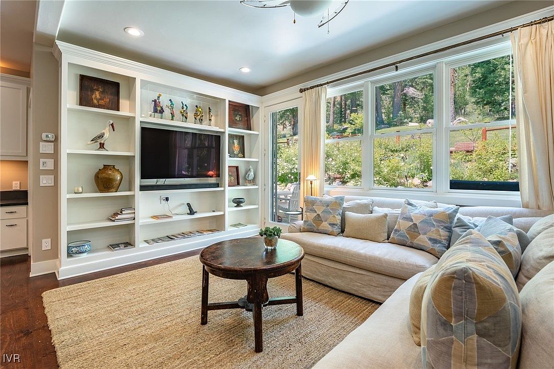 This is an interior shot of a cozy living room featuring a built-in bookshelf with a television, a round wooden coffee table, and a comfortable sofa with decorative pillows. Large windows provide ample natural light and a view of the outdoors, enhancing the room's inviting atmosphere. The room is decorated in neutral tones with wood accents.