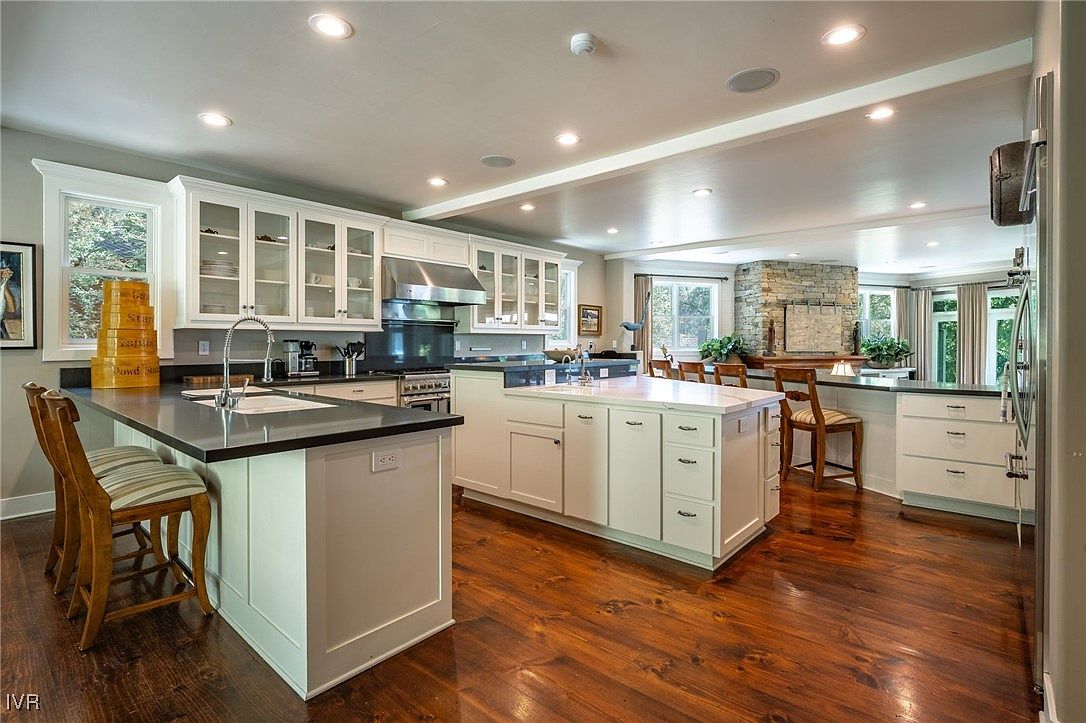 This is a spacious kitchen featuring white cabinetry, stainless steel appliances, and dark countertops on the island and perimeter counters. The hardwood flooring adds warmth to the space, and the open layout connects the kitchen to the dining area. The kitchen has two islands, one with a sink and the other with a cooktop.