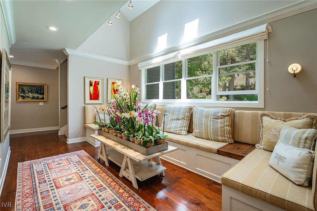 This interior shot showcases a charming hallway featuring hardwood floors and a cozy window seat. The window seat is adorned with striped cushions and decorative pillows, creating an inviting nook. A rustic wooden bench displays potted orchids, adding a touch of nature to the space, while artwork adorns the walls, enhancing the home's character.