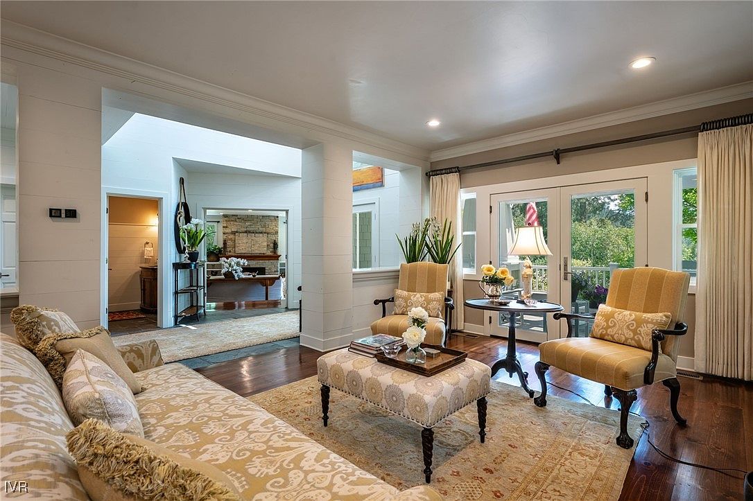 This is an interior shot of a living room, showcasing a comfortable and inviting space. The room features a patterned sofa, two armchairs, and an ottoman, all arranged on a patterned rug. The room opens to another space with a fireplace, creating an open and airy feel.
