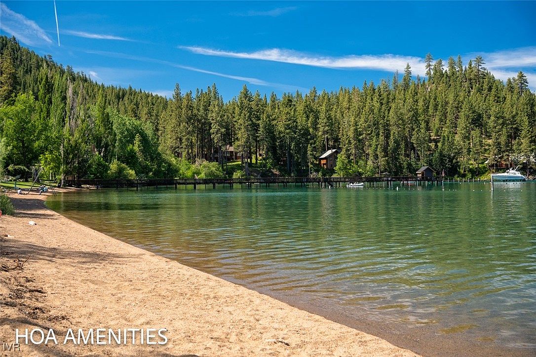 The image showcases a serene lakeside amenity, featuring a sandy beach in the foreground that transitions into clear, turquoise water. A wooden pier extends into the lake, connecting to small structures nestled among the lush green trees lining the shore. The scene is bathed in sunlight under a clear blue sky, creating an inviting and picturesque community space.