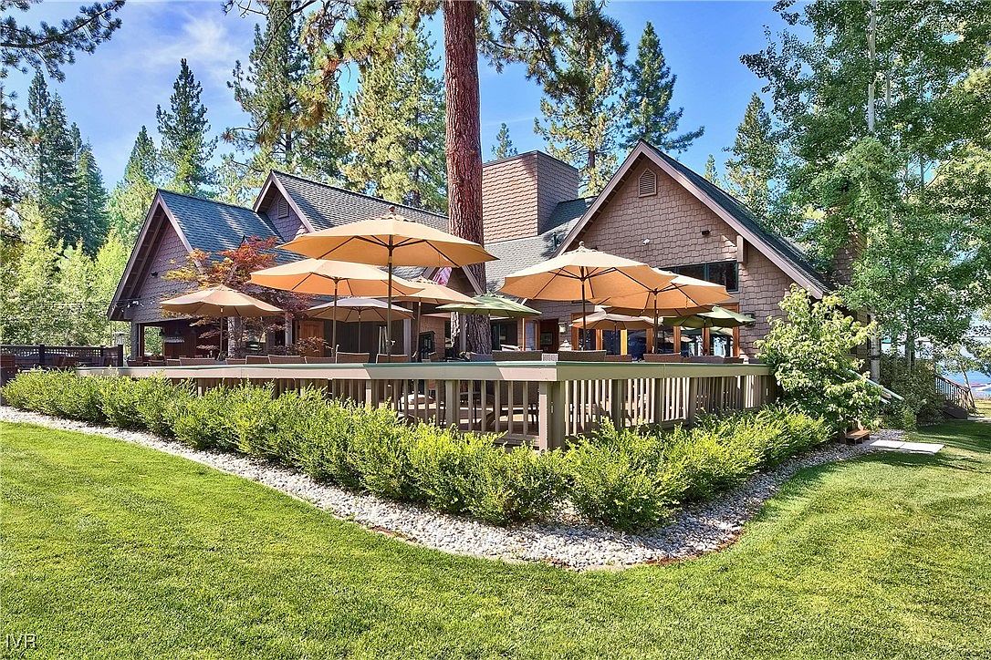 This image showcases a spacious deck area attached to a charming house, surrounded by lush greenery and mature trees. The deck is furnished with multiple patio umbrellas, suggesting an ideal space for outdoor dining and relaxation. The well-maintained lawn and landscaping add to the property's appeal, creating a serene and inviting atmosphere.
