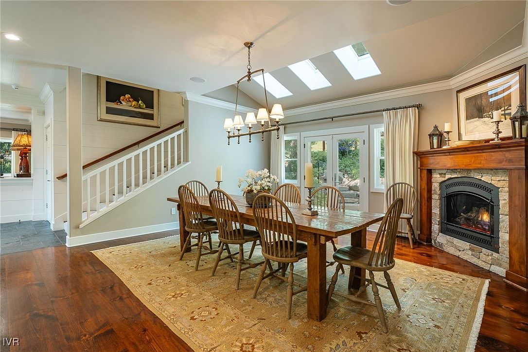 This is an inviting dining room featuring a large wooden table surrounded by Windsor-style chairs, set upon a patterned area rug. A stone fireplace with a wooden mantel adds warmth and character to the space, while a chandelier provides overhead lighting. Natural light streams in through skylights and a set of French doors, creating a bright and welcoming atmosphere.