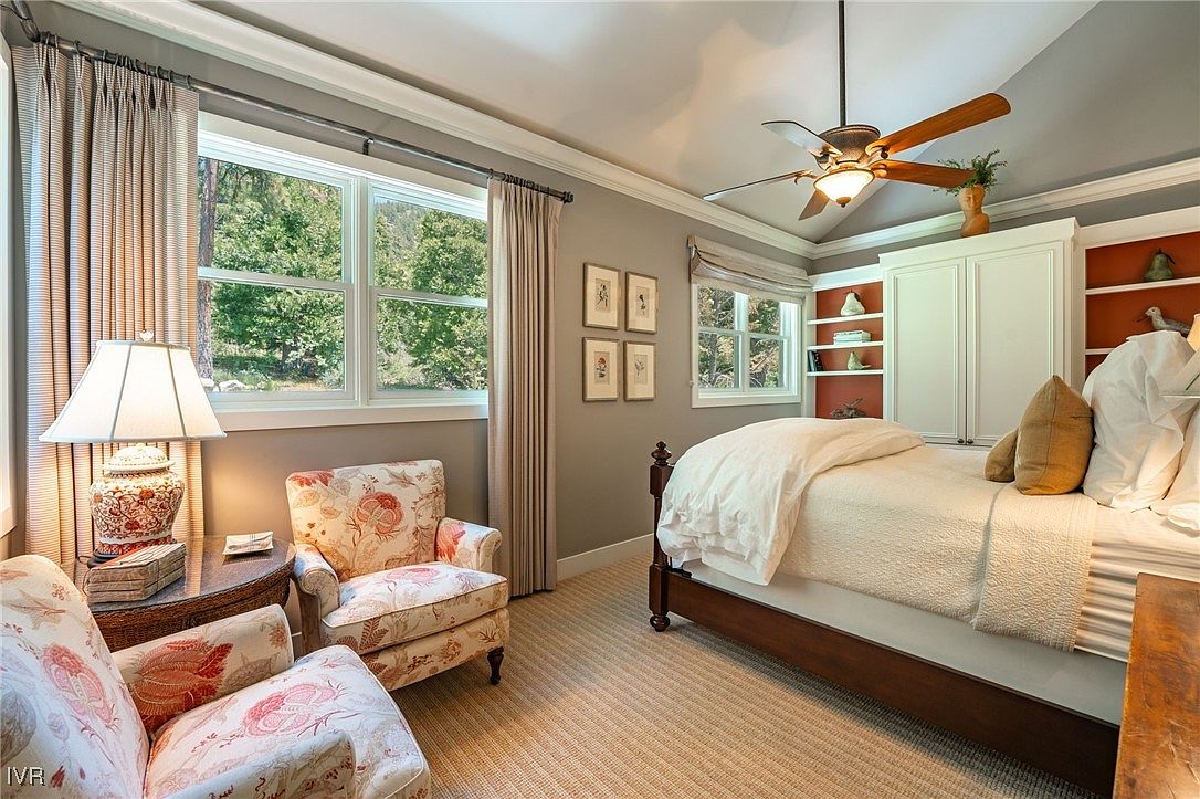 This is a cozy primary bedroom featuring a bed with a white comforter and several pillows. There are two floral armchairs near a window with curtains, and a ceiling fan hangs above the bed. Built-in shelving and cabinets provide ample storage, and the overall style is traditional and inviting.