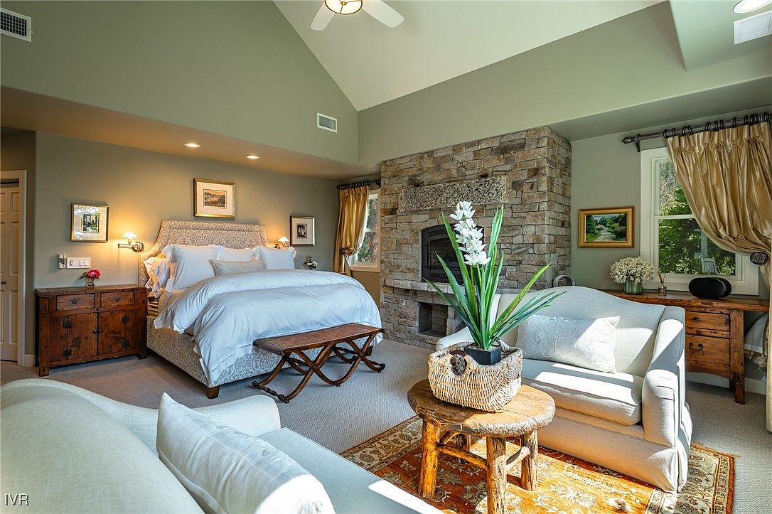 This is a well-lit primary bedroom featuring a king-size bed with white linens, a stone fireplace, and comfortable seating. The room is decorated in neutral tones with wooden accents, creating a warm and inviting atmosphere. The perspective is from the front of the room, showcasing the bed, fireplace, and seating area.