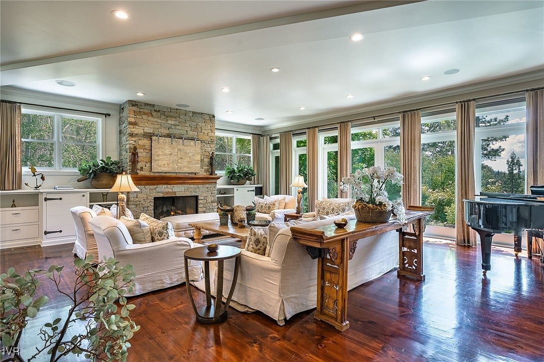 This is a spacious living room featuring hardwood floors, a stone fireplace, and large windows that provide ample natural light. The room is furnished with comfortable sofas, a wooden coffee table, and a console table behind the sofa, creating a cozy and inviting atmosphere. A grand piano sits near the windows, adding a touch of elegance.