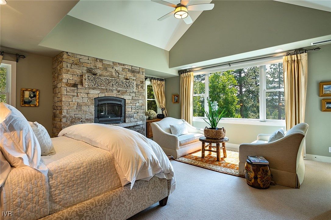 This is a primary bedroom featuring a stone fireplace, a large window with natural light, and two comfortable armchairs. The room is decorated in neutral tones with light-colored walls and carpet, creating a serene and inviting atmosphere. The bed is neatly made with white linens, and the overall impression is one of luxury and comfort.