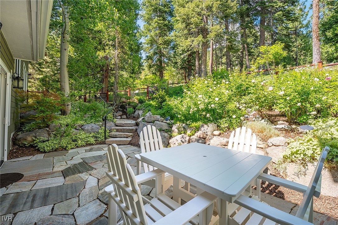 This image showcases a charming outdoor patio area, featuring a white wooden table and chairs set on a stone-tiled surface. The patio is surrounded by lush greenery, including trees, bushes, and flowering plants, creating a serene and private atmosphere. Stone steps lead up to a higher level of the garden, adding visual interest and depth to the outdoor space.