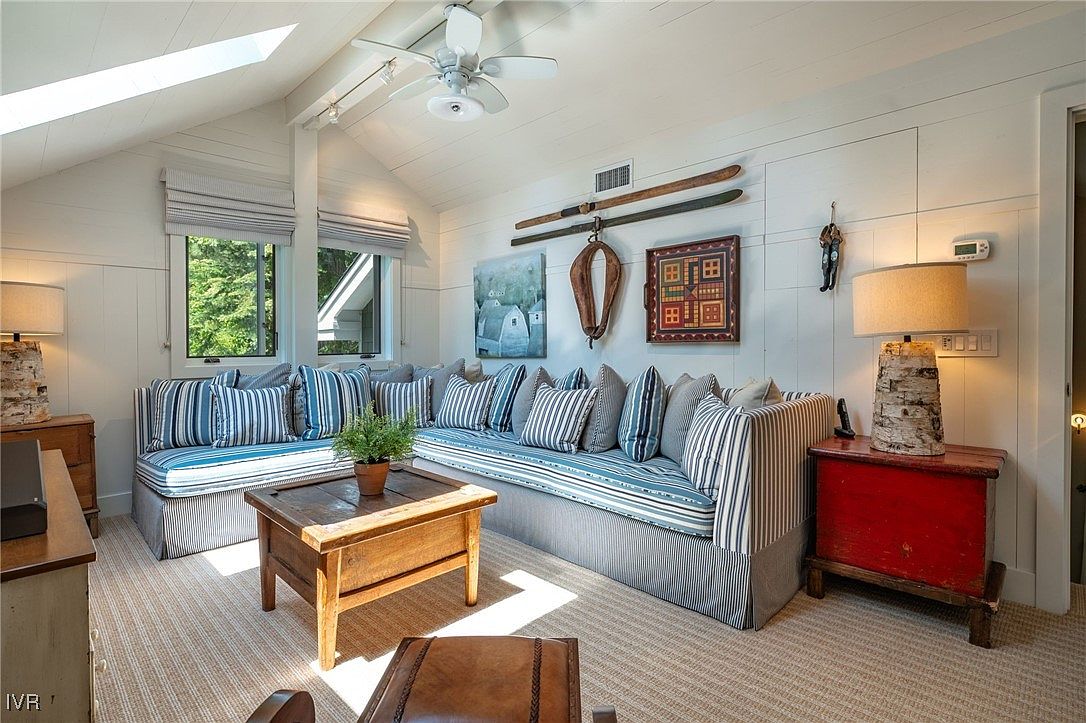 This is a cozy living room featuring a large, sectional sofa with blue and white striped cushions and a wooden coffee table. The room has a vaulted ceiling with a skylight, and the walls are decorated with artwork and vintage skis. A red side table with a birch lamp adds a pop of color, creating a warm and inviting atmosphere.