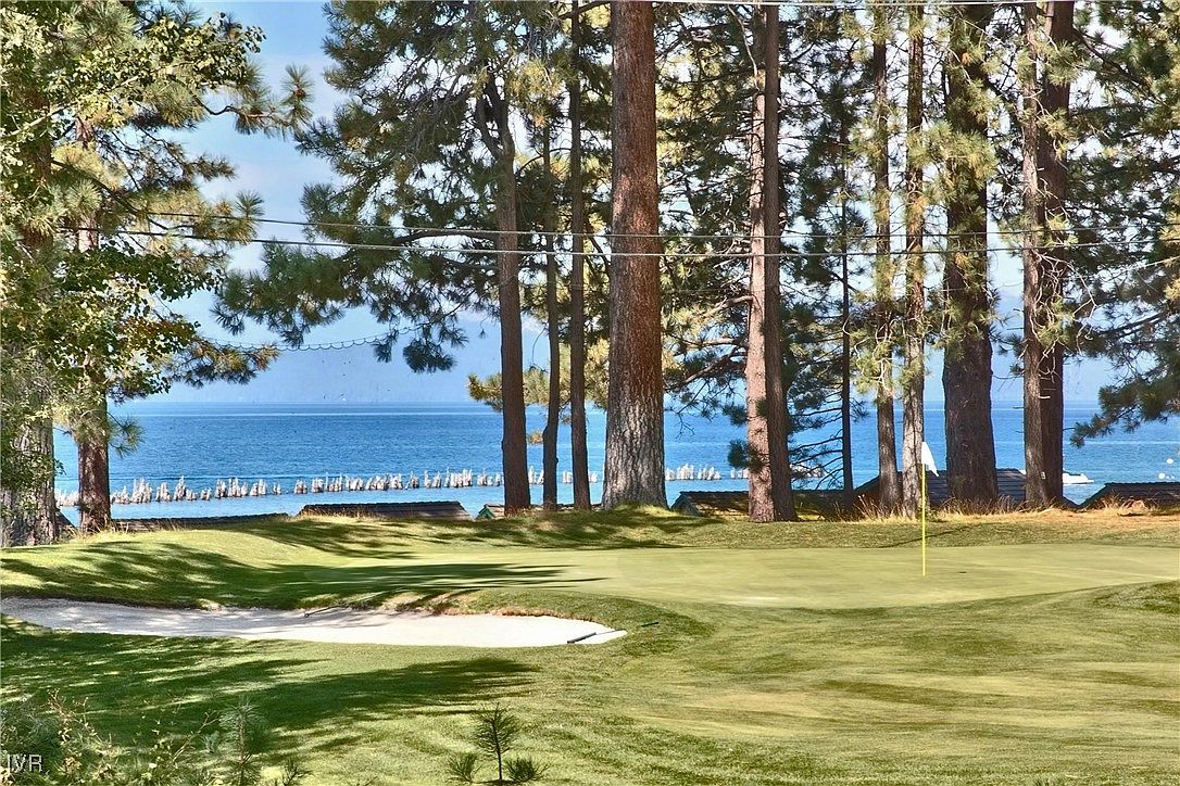 This image showcases a beautifully maintained golf course with a view of a serene lake in the background. Tall trees frame the scene, adding a sense of tranquility and privacy. A sand trap is visible in the foreground, and a flag marks the green, highlighting the course's features.