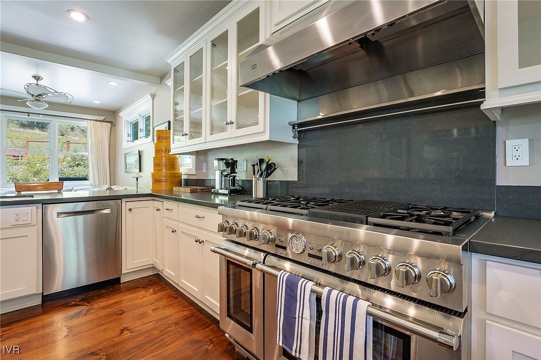 This is a well-appointed kitchen featuring white cabinetry, dark countertops, and stainless steel appliances. A large stainless steel range with a prominent vent hood is the focal point, complemented by glass-front cabinets above the countertop. The warm wood flooring adds a touch of traditional charm to the space.
