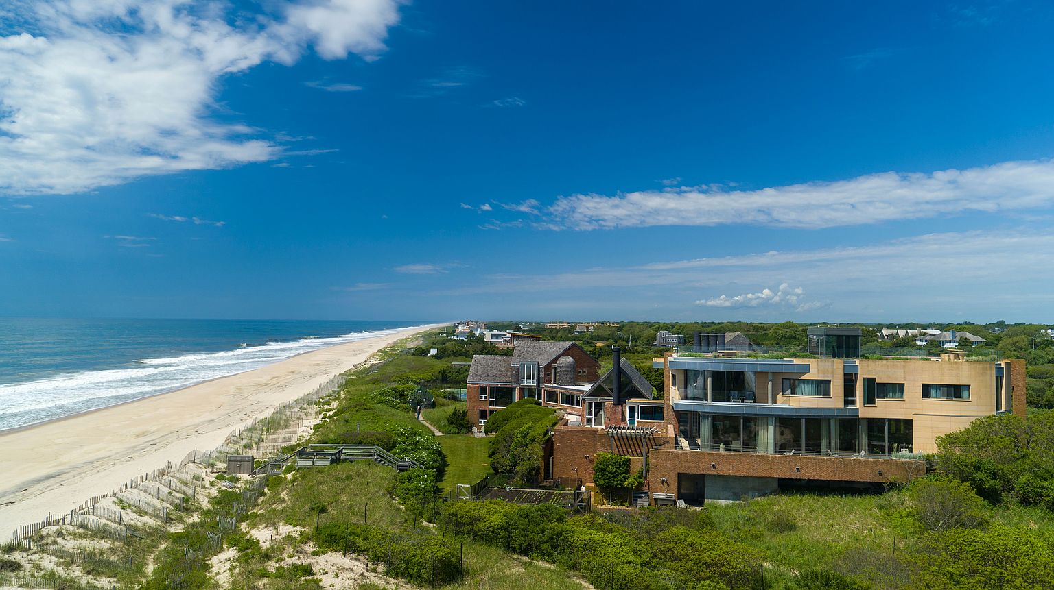 This aerial view showcases a stunning beachfront property featuring modern architectural design with a mix of brick and glass elements. The property is nestled amidst lush greenery, offering privacy and a seamless blend with the natural landscape. The expansive sandy beach and ocean provide a picturesque backdrop, emphasizing the property's prime location and luxurious appeal.