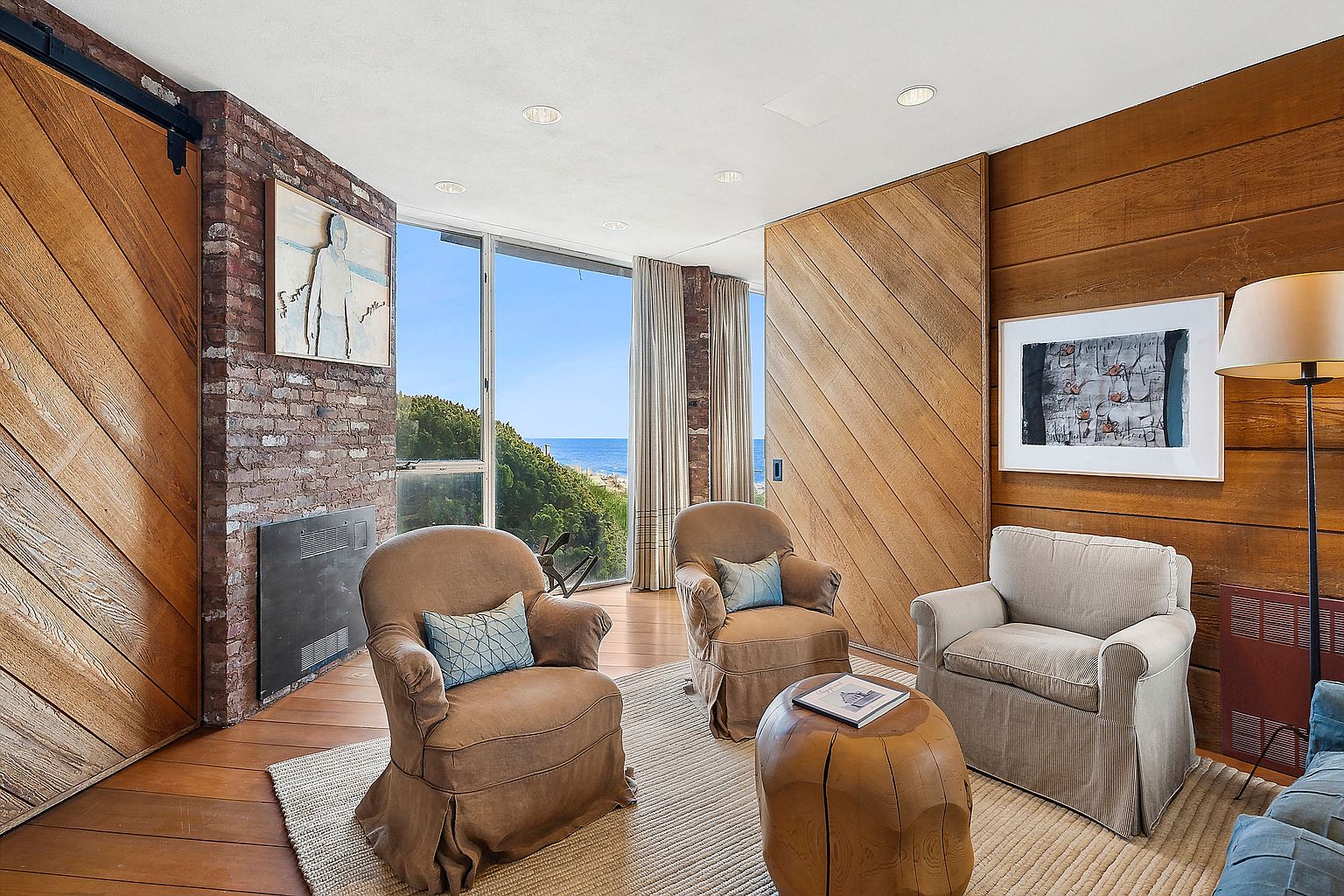 This inviting living room features a blend of rustic and modern elements, with exposed brick and wood paneling creating a warm atmosphere. Three comfortable armchairs are arranged around a unique wooden coffee table, set upon a neutral-toned rug. Large windows offer a glimpse of the ocean, enhancing the room's serene and relaxing ambiance.