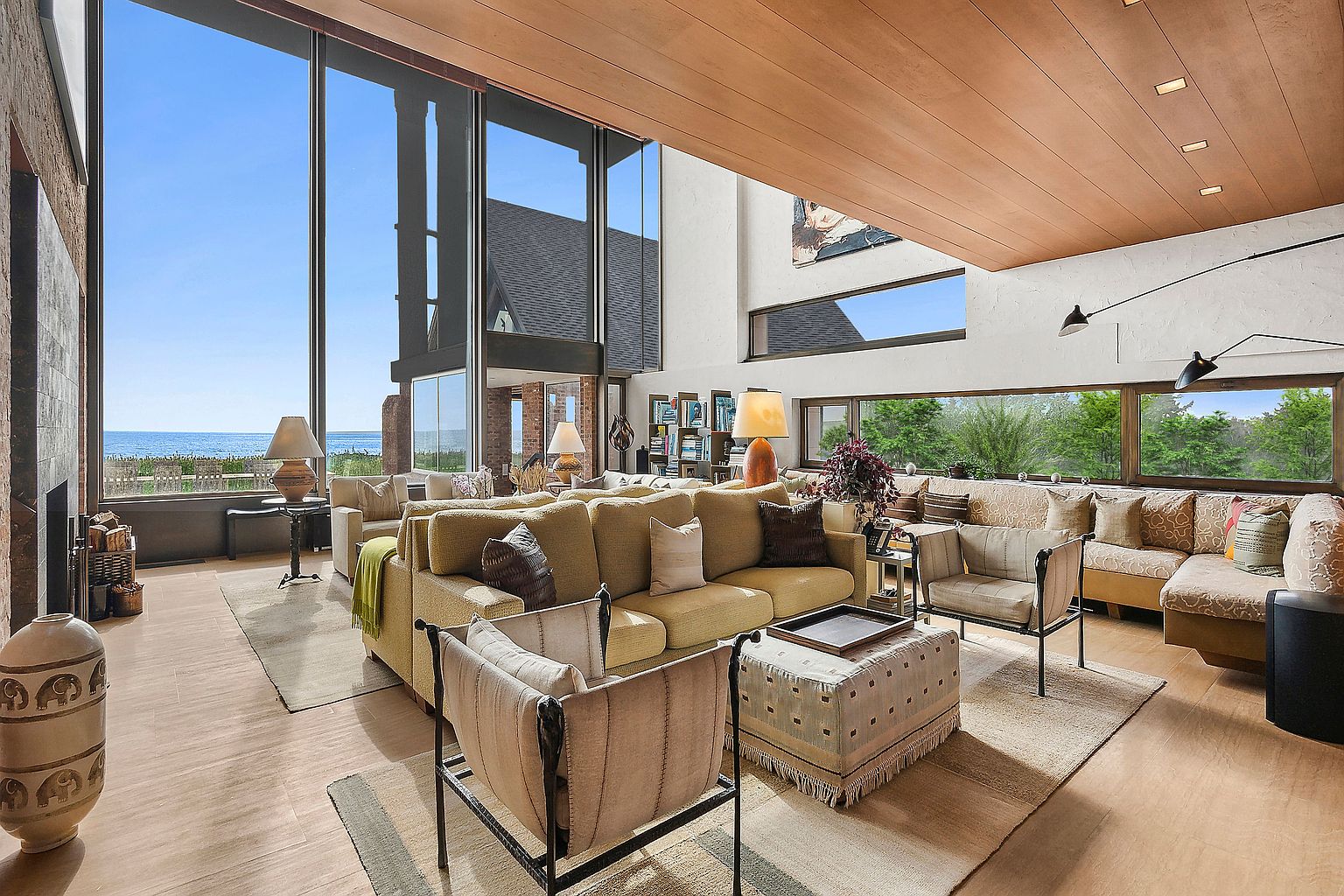 This is a spacious living room with floor-to-ceiling windows offering ocean views. The room features a large sectional sofa, comfortable armchairs, and a neutral color palette, creating a relaxing and inviting atmosphere. The wood-paneled ceiling and unique architectural details add character and sophistication to the space.