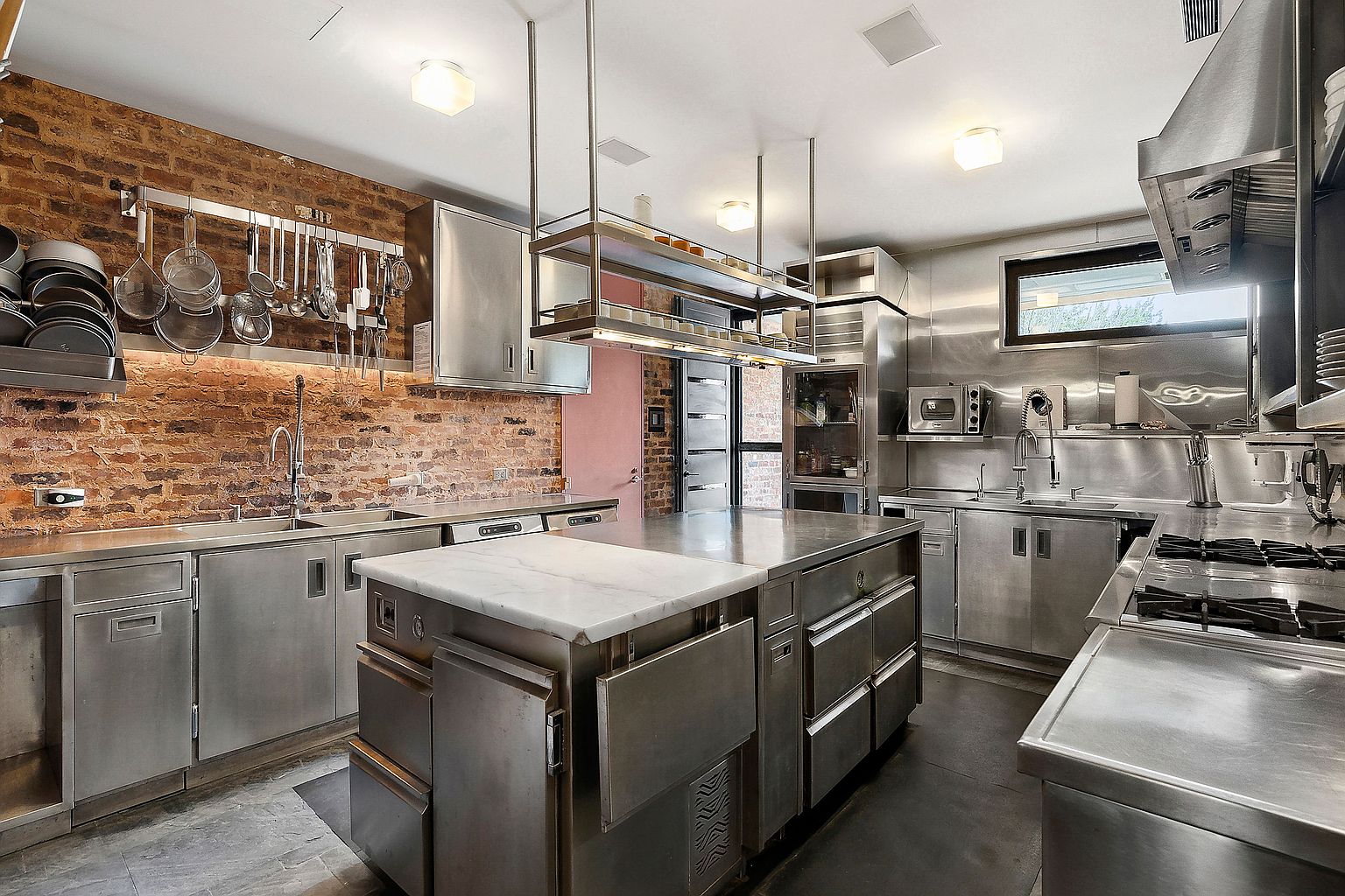 This is a professional-grade kitchen featuring stainless steel cabinetry, appliances, and countertops, accented by a brick wall. A large island with a marble top stands centrally, with a pot rack hanging above. The space is well-lit and designed for functionality and durability.