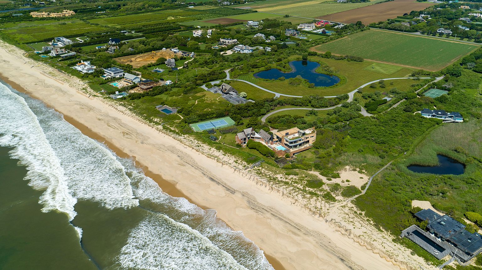 This aerial shot showcases a luxurious beachfront property with a sprawling lawn, tennis court, and a modern architectural design. The property is nestled amidst lush greenery and features a private beach access, offering stunning ocean views. The overall impression is one of exclusivity and high-end coastal living.