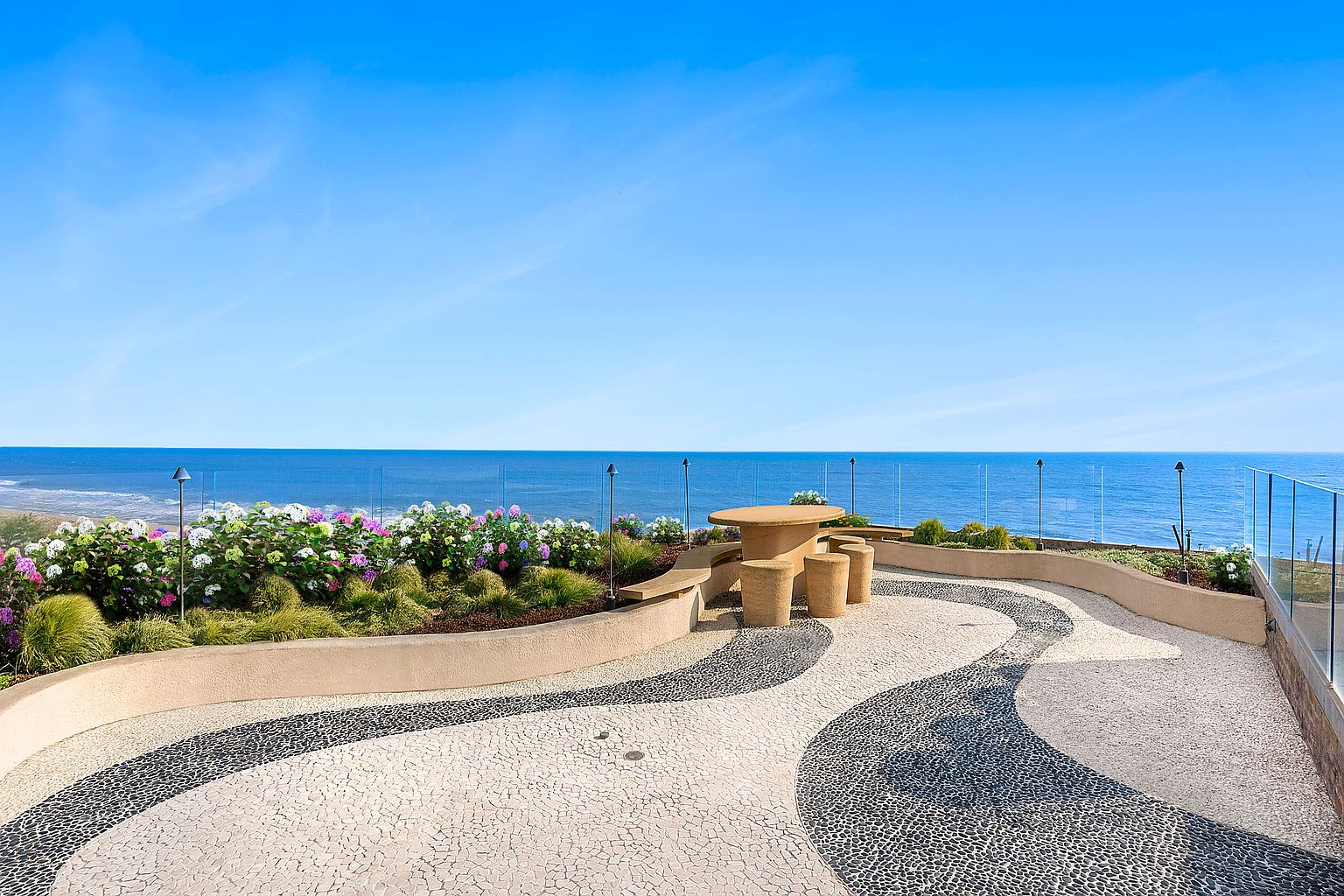 This image showcases a beautifully designed patio or deck area with a stunning ocean view. The space features a unique patterned stone walkway, a built-in seating area with a table, and lush landscaping with colorful flowers. A glass railing provides an unobstructed view of the ocean, enhancing the property's appeal and creating a serene outdoor living space.