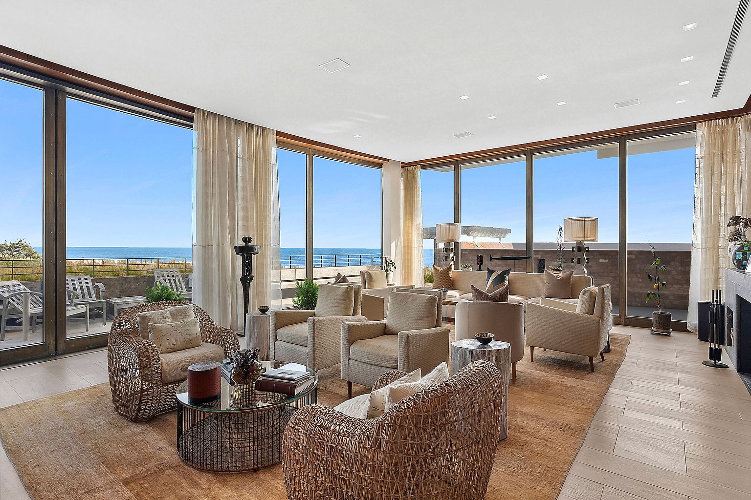 This is a luxurious living room with floor-to-ceiling windows offering stunning ocean views. The room features comfortable seating arrangements with wicker and upholstered armchairs, a glass-topped coffee table, and a neutral color palette. The overall impression is one of elegance and relaxation, perfect for enjoying the coastal scenery.