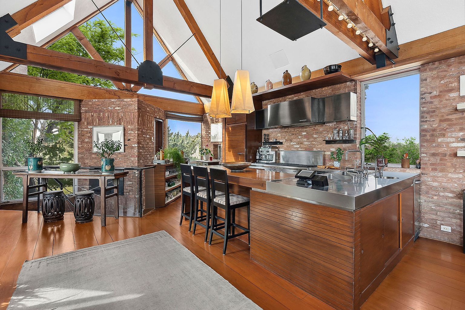 This is an interior shot of a spacious kitchen featuring a large island with a stainless steel countertop and wooden base. The kitchen has brick walls, wooden beams, and large windows that provide natural light. The overall style is modern with rustic elements, creating a warm and inviting atmosphere.