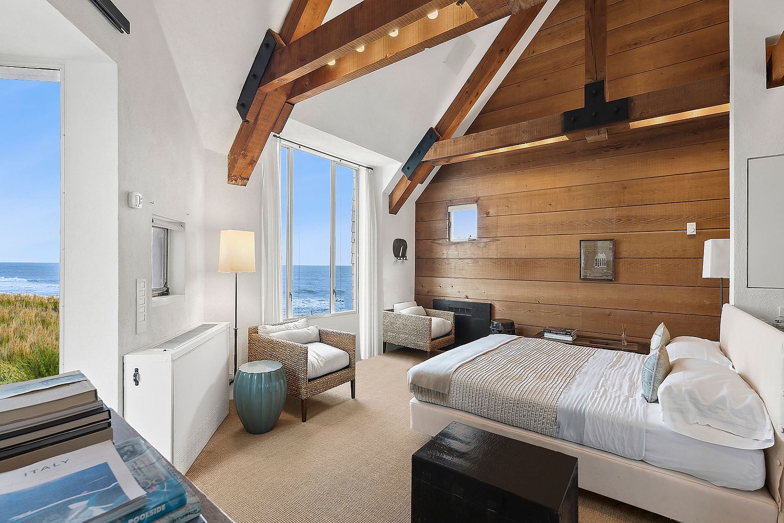This primary bedroom features a vaulted ceiling with exposed wooden beams and a wood-paneled accent wall. Large windows offer ocean views, and the room is furnished with a bed, two wicker chairs, and a unique blue side table. The overall aesthetic is rustic and coastal, creating a serene and inviting space.