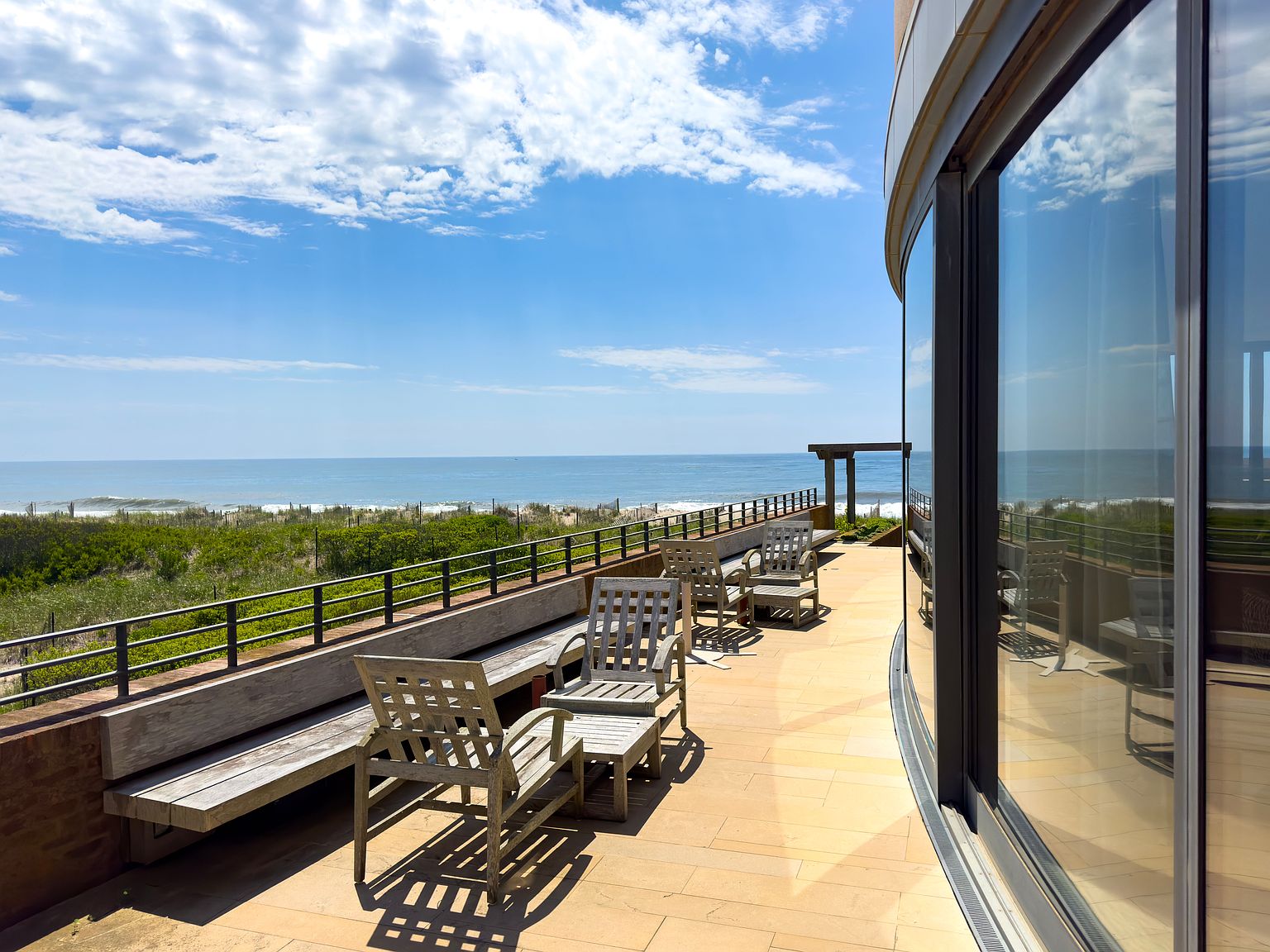 This image showcases a luxurious patio or balcony with stunning ocean views. The space is furnished with wooden chairs and benches, creating a relaxing outdoor living area. A sleek railing and modern architectural details add to the upscale feel, making it an ideal spot for enjoying the coastal scenery.