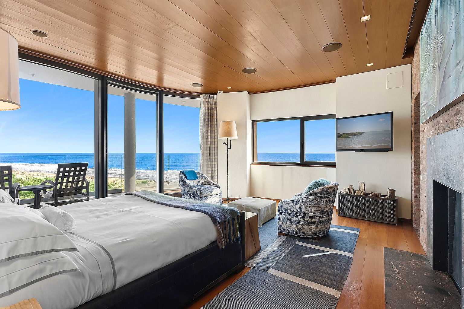 This is a luxurious primary bedroom with stunning ocean views through large windows. The room features a king-size bed, two armchairs, a fireplace, and a flat-screen TV. The wood-paneled ceiling adds warmth, while the blue and gray rug complements the coastal aesthetic.