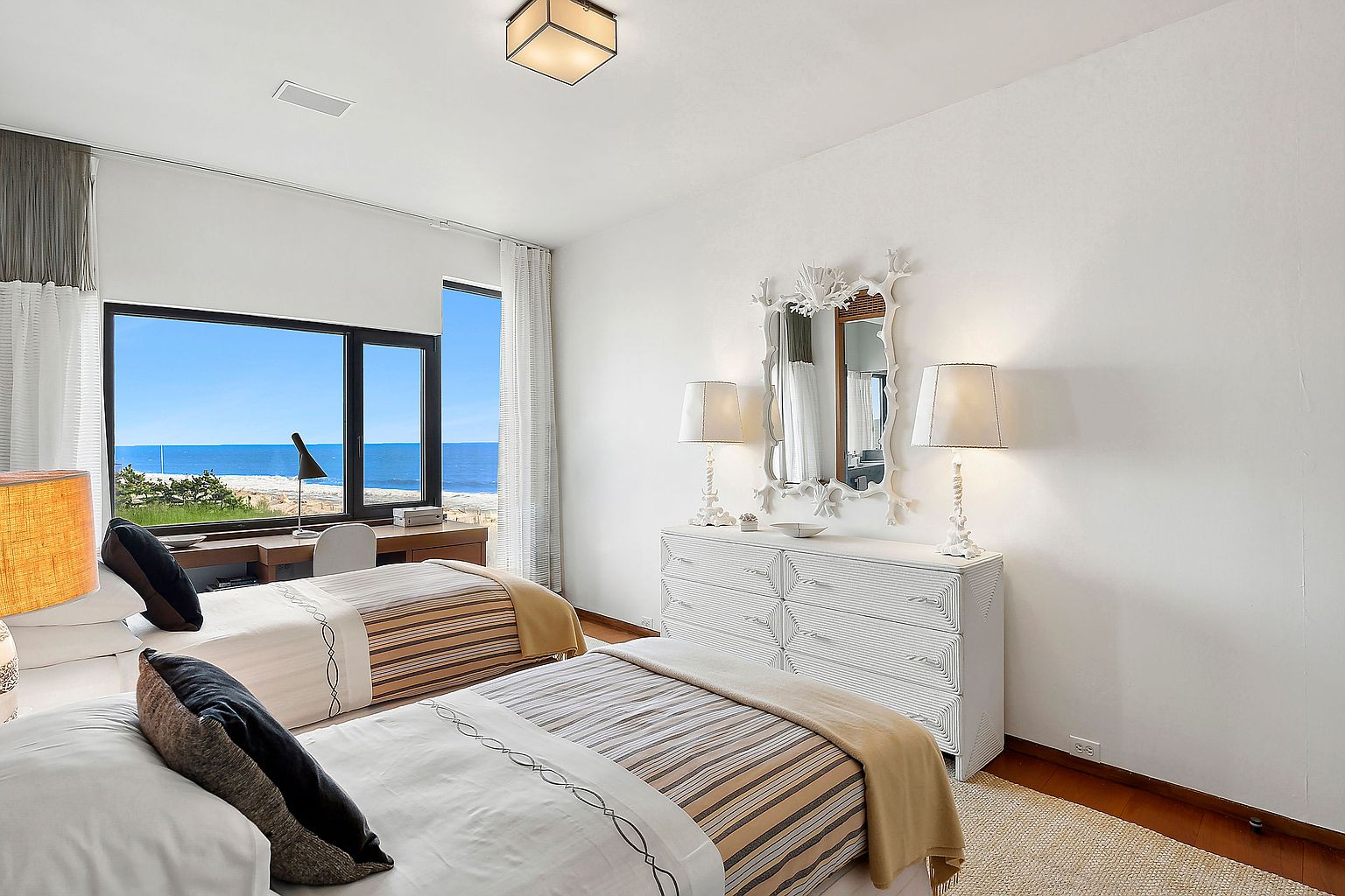 This is a bright and airy guest bedroom featuring two twin beds with striped bedding and a neutral color palette. A large window offers a stunning ocean view, complemented by a built-in desk area. The room also includes a white dresser with decorative lamps and a mirror, enhancing the coastal chic aesthetic.