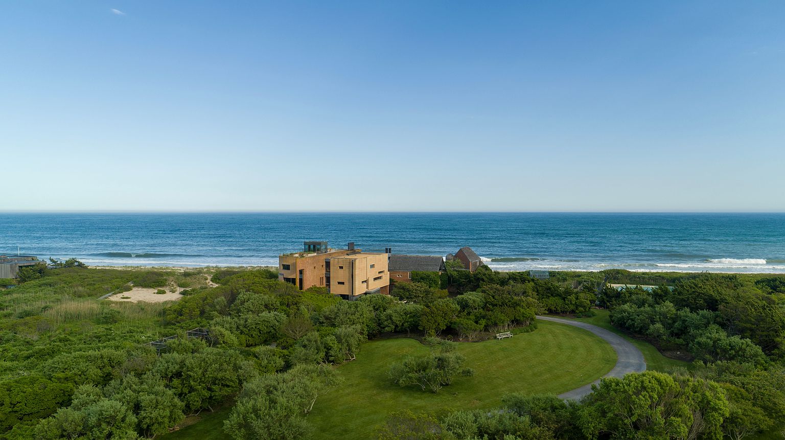 This aerial shot showcases a luxurious beachfront property with a modern architectural design. The house is surrounded by lush greenery and features a winding driveway leading to the entrance. The property offers stunning ocean views and a sense of privacy and exclusivity.