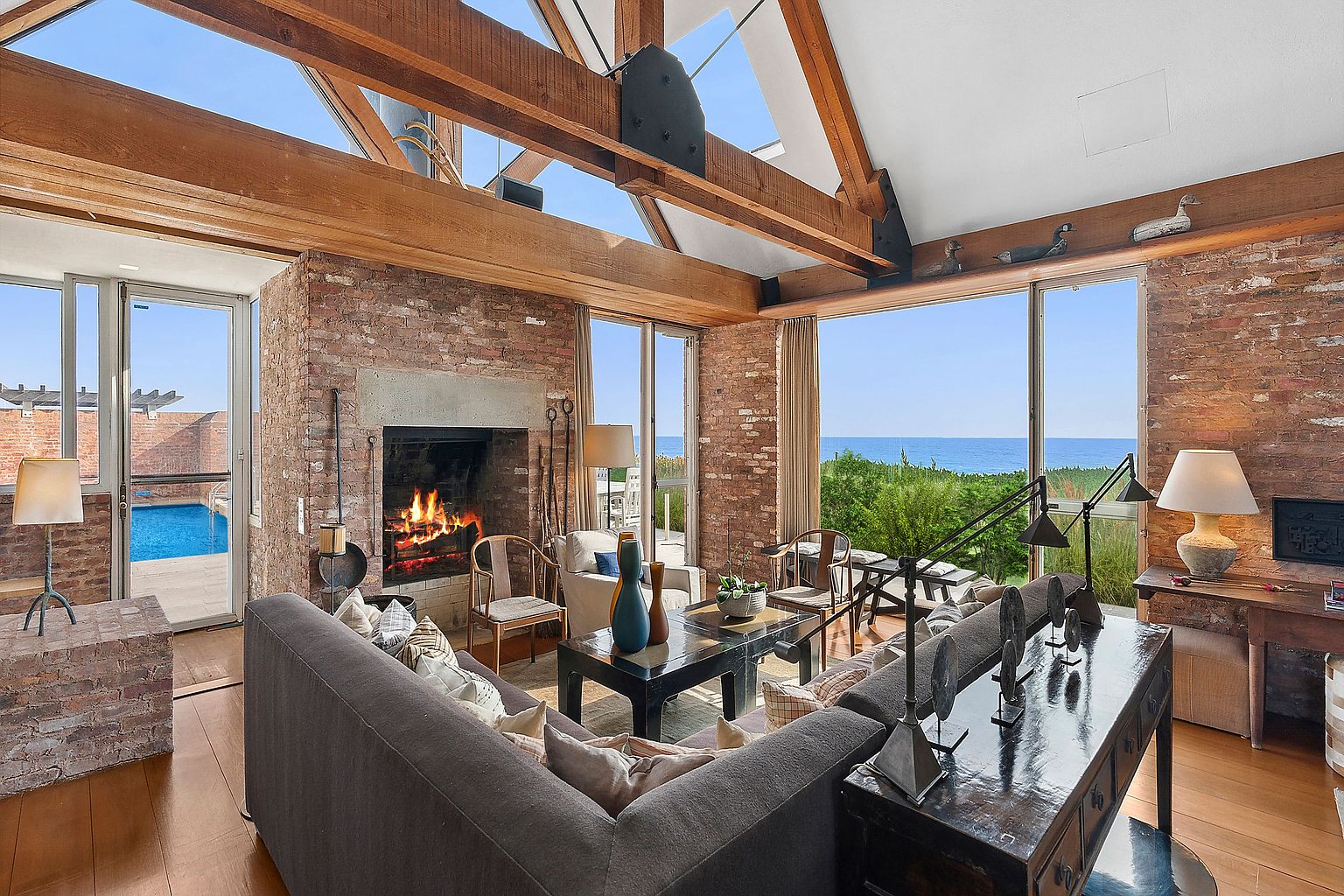 This is an interior shot of a living room featuring a brick fireplace, wooden beams, and large windows with ocean views. The room is furnished with a comfortable gray sofa, a dark wood coffee table, and various decorative items. The overall style is rustic and luxurious, creating a warm and inviting atmosphere.