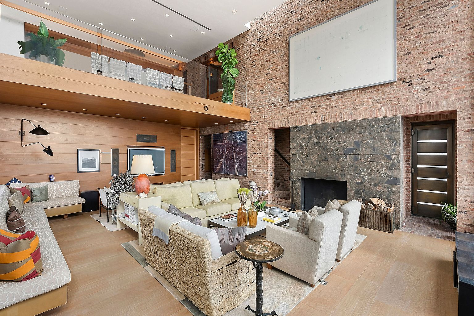 This is a spacious living room featuring a mix of textures and materials, including exposed brick, wood paneling, and various seating options. The room is well-lit and includes a fireplace, a large blank canvas, and a mezzanine level, creating a unique and inviting atmosphere. The overall impression is one of modern comfort and artistic flair.