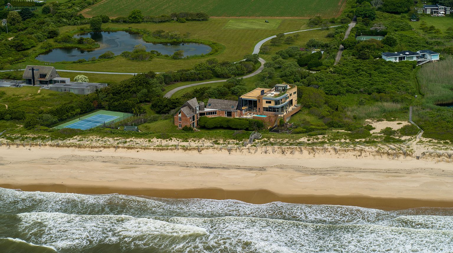 This aerial view showcases a luxurious beachfront property with a modern architectural design. The residence features a combination of brick and contemporary elements, including a multi-level structure with a pool and direct beach access. The surrounding landscape includes lush greenery, a tennis court, and a serene pond, enhancing the property's exclusivity and appeal.