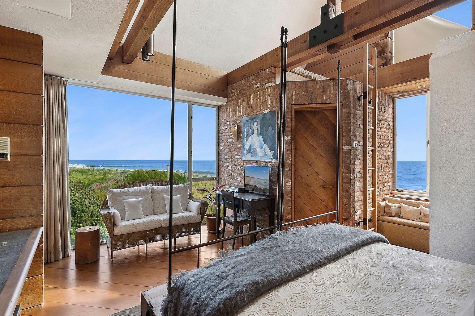 This primary bedroom features a unique blend of rustic and coastal elements. Exposed brick walls and wooden beams create a warm, textured backdrop, while large windows offer stunning ocean views. A four-poster bed with a cozy throw adds to the inviting atmosphere, complemented by a small desk area and comfortable seating.