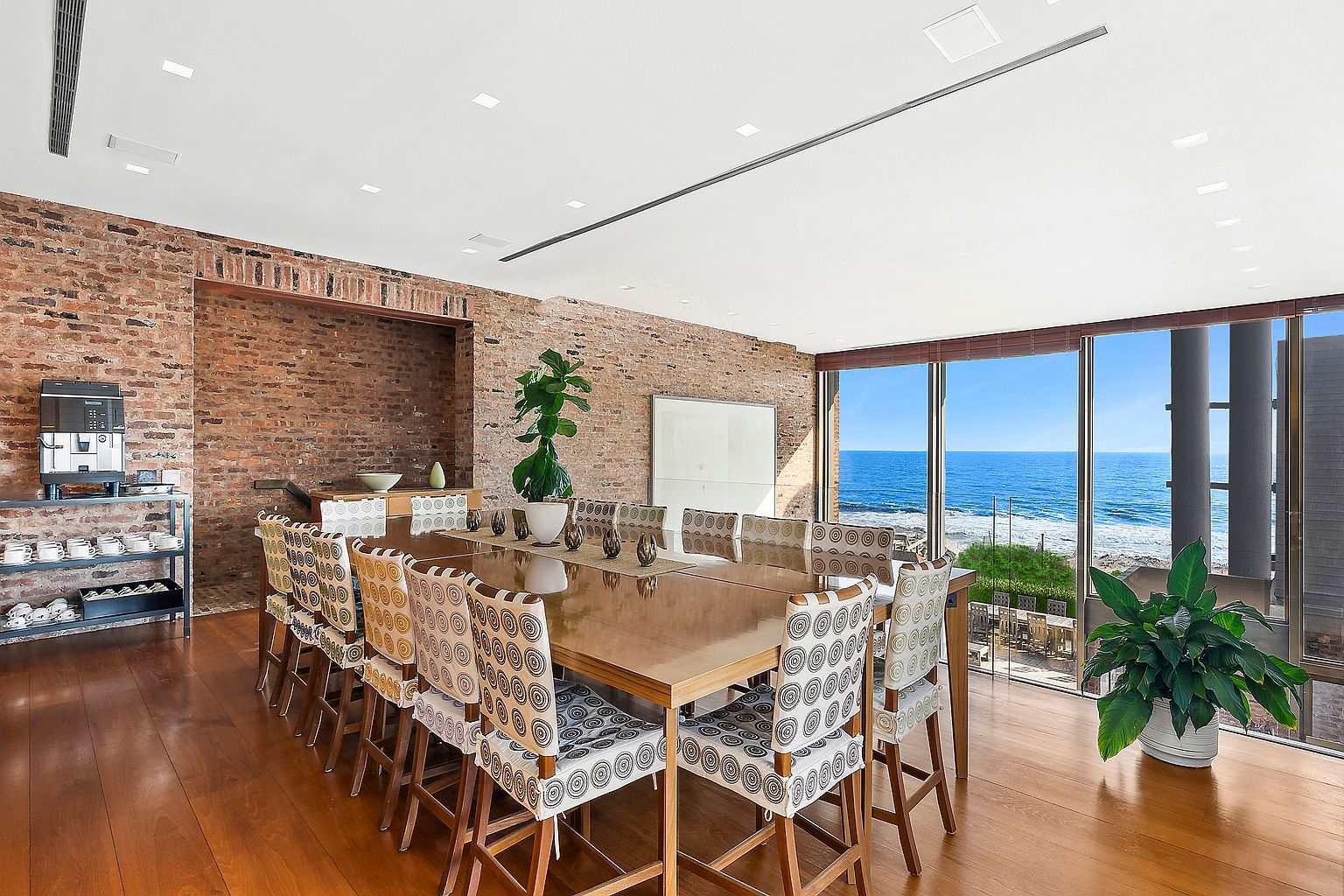 This is a dining room with a large wooden table surrounded by patterned chairs. The room features a brick wall and large windows offering an ocean view. There is a coffee machine on a metal shelf and a large plant in the corner, adding a touch of nature to the space.