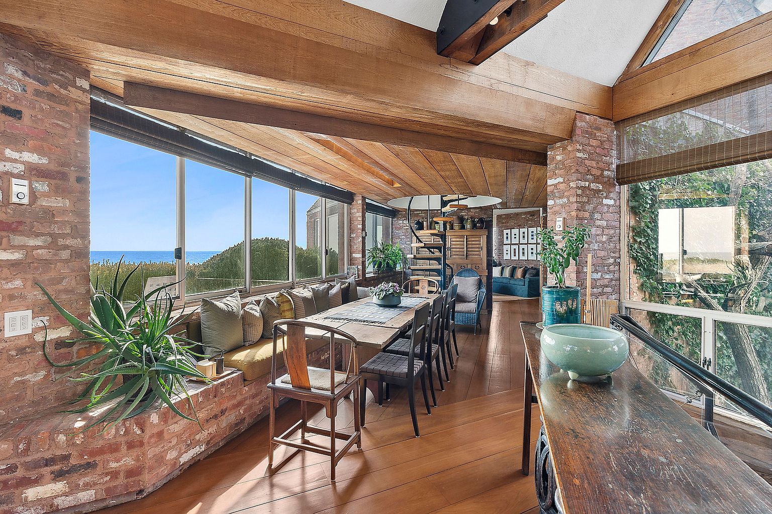 This is an interior shot of a dining room featuring a long wooden table with chairs, set against a backdrop of large windows offering ocean views. The room is characterized by its wooden floors, exposed brick accents, and a unique spiral staircase in the background. The overall impression is one of rustic elegance and spaciousness, enhanced by natural light and scenic views.