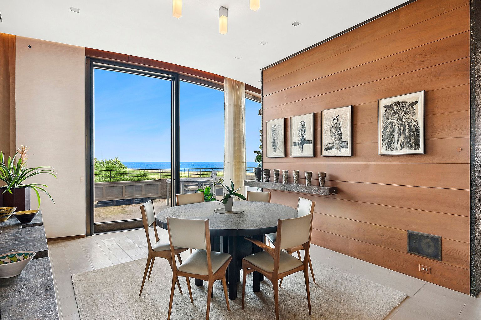 This dining room features a round table with six chairs, set on a light-colored rug. A large window offers a view of the ocean, and the wall is decorated with artwork and a shelf holding decorative items. The room has a modern and elegant feel, with natural light enhancing the space.