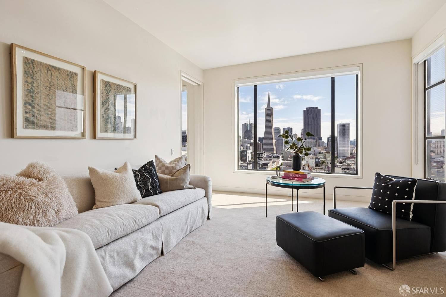 This is an interior shot of a living room featuring a comfortable sofa with decorative pillows, a modern black chair and ottoman, and a round glass-topped table. Large windows offer a stunning city view, enhancing the room's appeal. The neutral color palette and artwork create a sophisticated and inviting atmosphere.