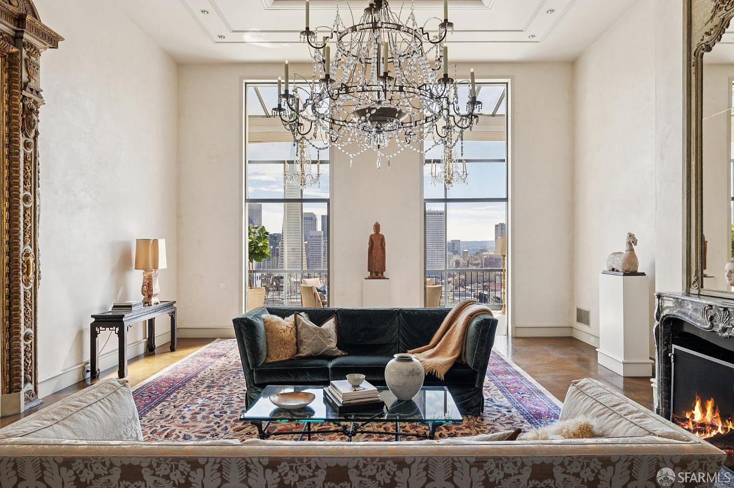 This is a luxurious living room featuring a large chandelier, floor-to-ceiling windows with city views, and a sophisticated color palette. The room is furnished with a plush sofa, a glass coffee table, and a decorative fireplace, creating an elegant and inviting atmosphere. The hardwood floors and ornate details add to the room's upscale appeal.
