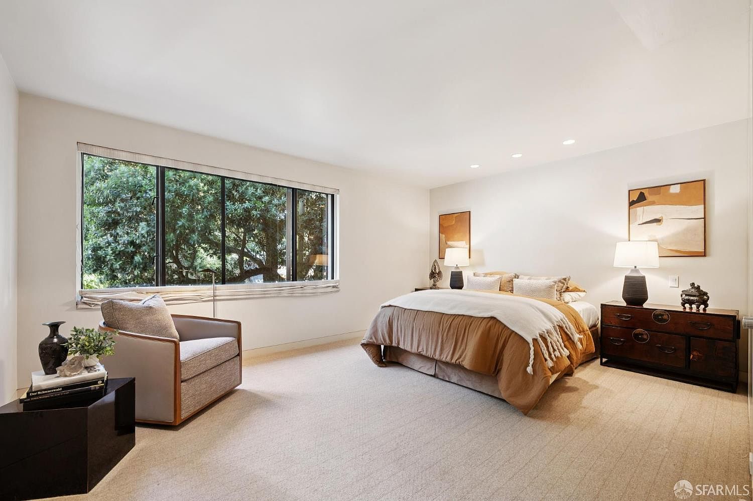 This is a well-lit primary bedroom featuring a large window with a view of lush greenery. The room is decorated in neutral tones, with a comfortable armchair, a bed with a tan comforter and white throw, and a dark wood dresser with lamps. The overall impression is one of calm and understated elegance.