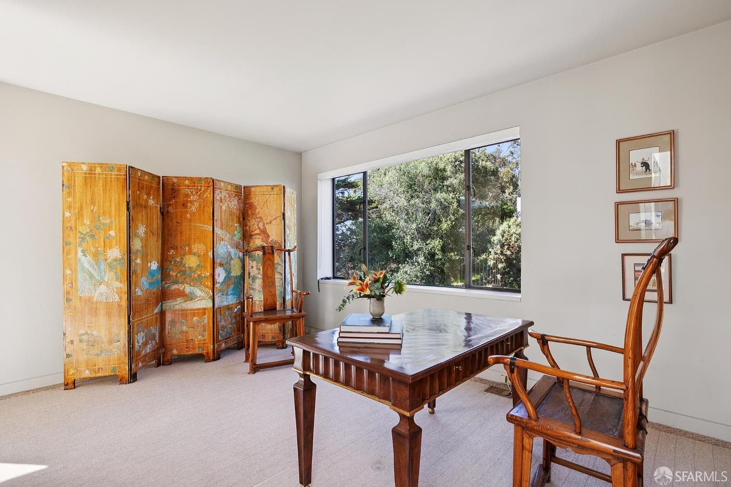 This interior shot showcases a well-lit office or study area. The room features a decorative folding screen, a wooden desk with books and flowers, and two wooden chairs. Natural light streams in through a large window, offering a view of lush greenery, while framed artwork adorns the wall, creating a serene and sophisticated workspace.