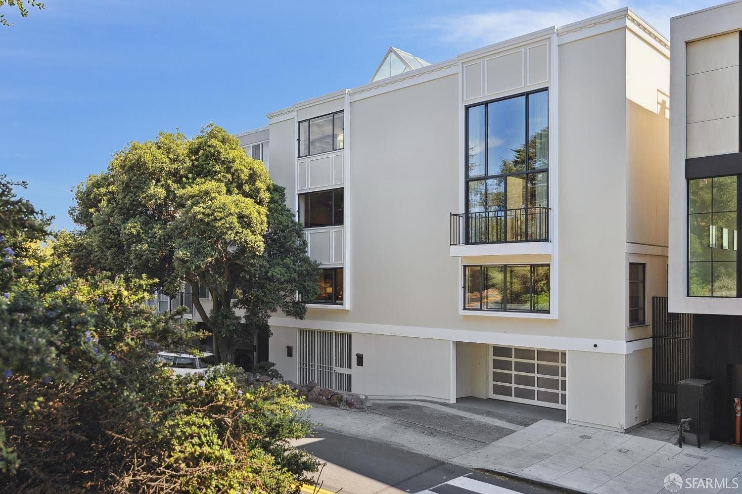This is a front exterior view of a modern multi-story building. The building features a light-colored facade, large windows with black frames, and a balcony. A mature tree partially obscures the view, adding a touch of nature to the urban setting. A garage door is visible on the ground floor.