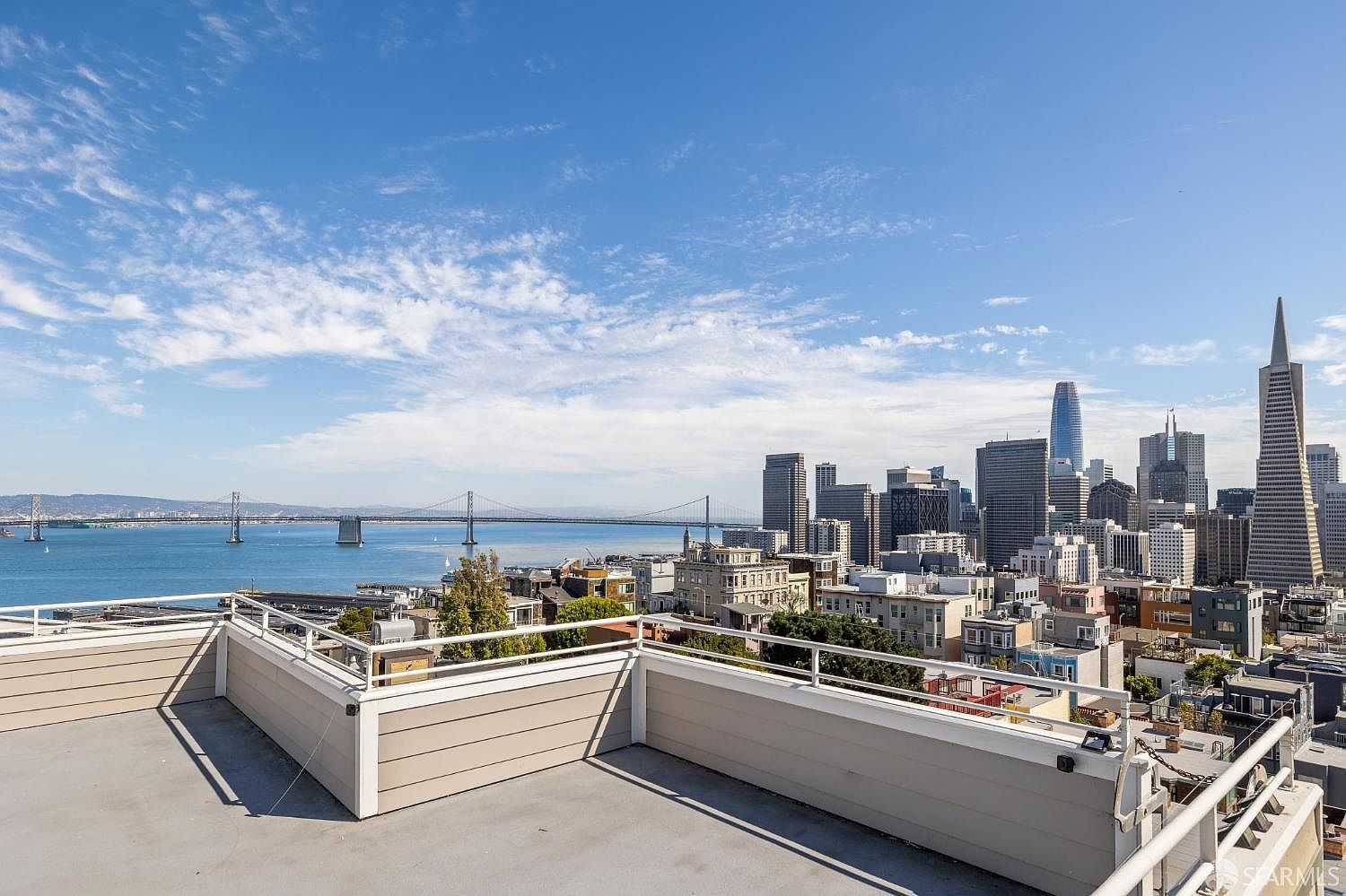 This image showcases a stunning rooftop patio/deck with panoramic views of the San Francisco skyline and the Bay Bridge. The deck features a modern railing system and a clean, minimalist design, offering an ideal outdoor space for relaxation and entertainment. The cityscape provides a dramatic backdrop, emphasizing the property's prime location and desirable urban lifestyle.