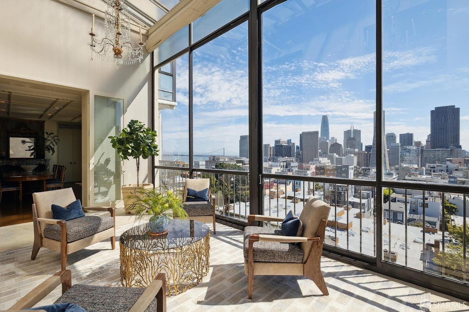 This sunroom features floor-to-ceiling windows offering a panoramic city view, including the Golden Gate Bridge. The room is furnished with comfortable armchairs arranged around a unique, decorative coffee table, creating an inviting space for relaxation. A potted tree adds a touch of nature to the interior.
