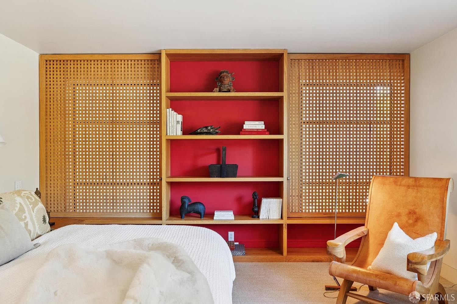 This is a bedroom featuring a bed with white linens, a wooden bookshelf with red backing, and a tan leather chair. The bookshelf is decorated with various objects and books. The room has a warm and inviting feel, with a focus on natural materials and textures.