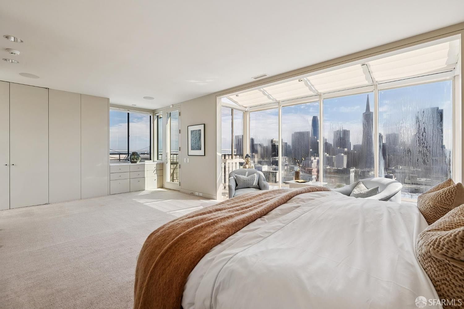 This is a bright and spacious primary bedroom featuring floor-to-ceiling windows with a stunning city view. The room includes a large bed with white linens and a brown throw, built-in wardrobes, and a seating area with comfortable chairs. The overall impression is luxurious and serene, emphasizing the view and ample space.