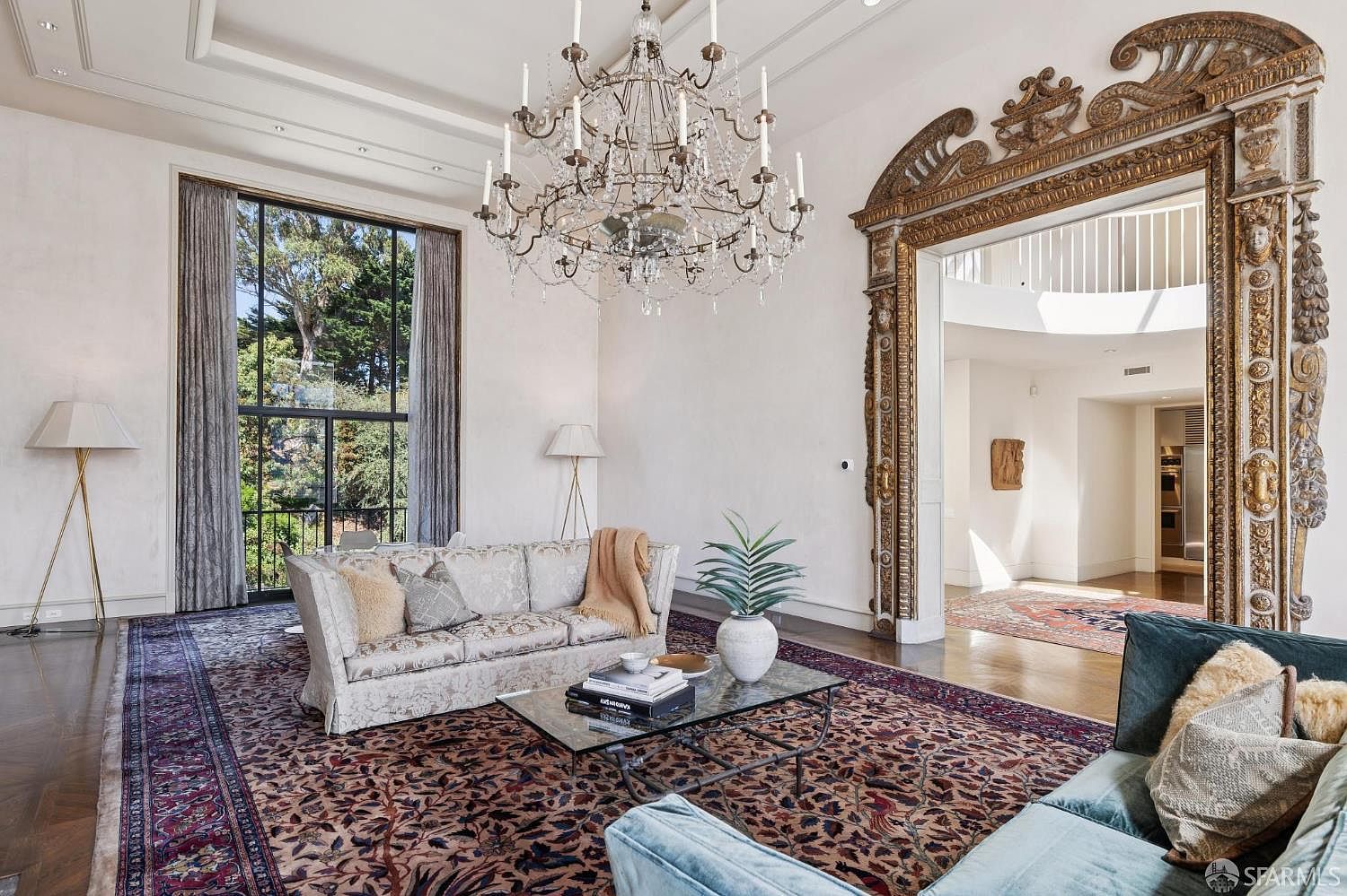 This is an interior shot of a luxurious living room featuring a large crystal chandelier, ornate doorway, and a patterned area rug. The room is furnished with a sofa and a glass coffee table, and natural light streams in through a large window, creating a bright and inviting atmosphere. The style is elegant and sophisticated, suggesting a high-end property.