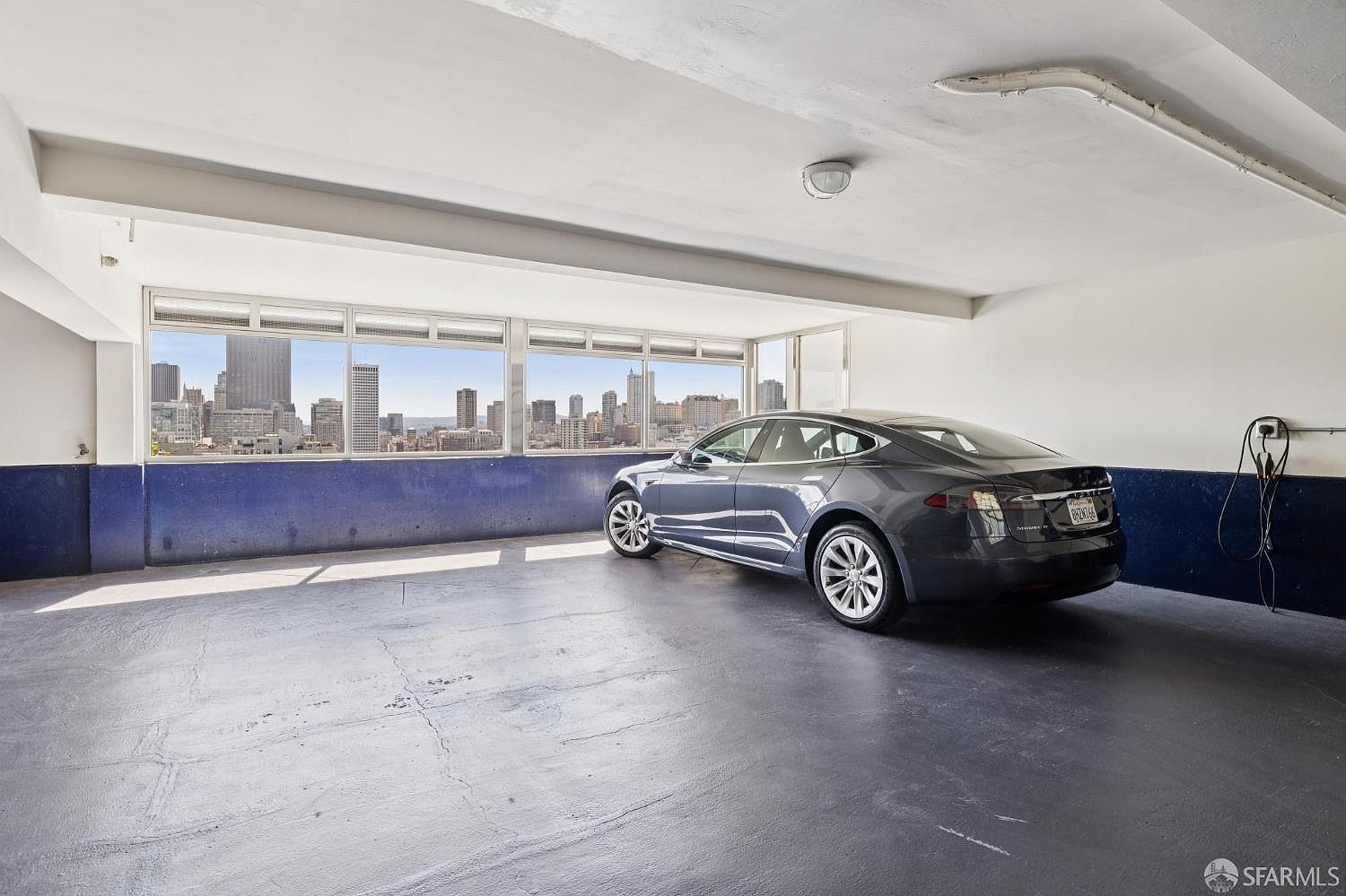 The image showcases a modern garage with a dark gray Tesla parked inside. The garage features a large window offering a stunning city view, a blue accent wall, and a charging station for the electric vehicle. The space is well-lit and appears clean, presenting a luxurious and functional area for vehicle storage and maintenance.